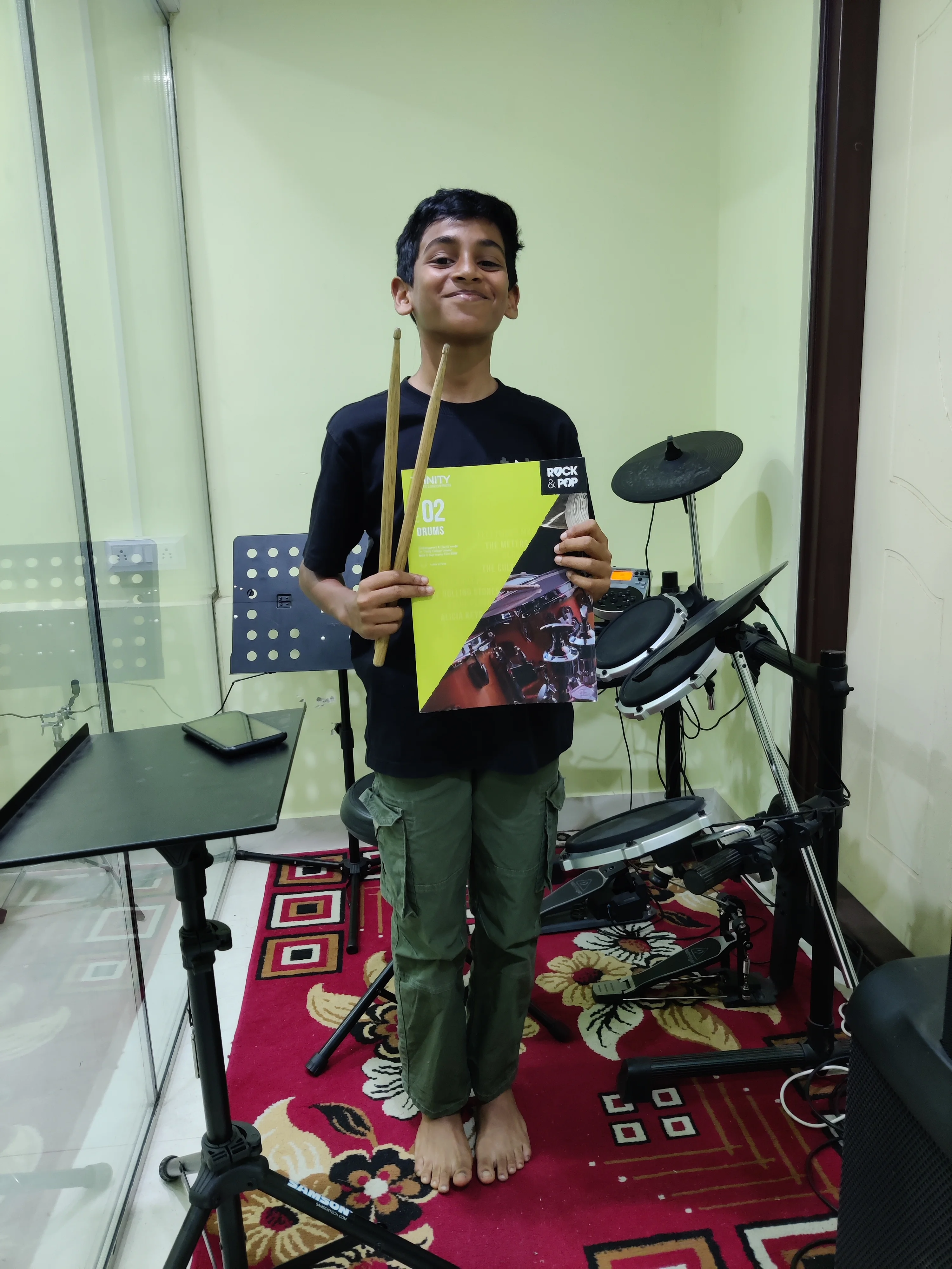 FSG drum student Aiden holding Trinity Rock and Pop Grade 2 Drums book with drumsticks — FSG practice room