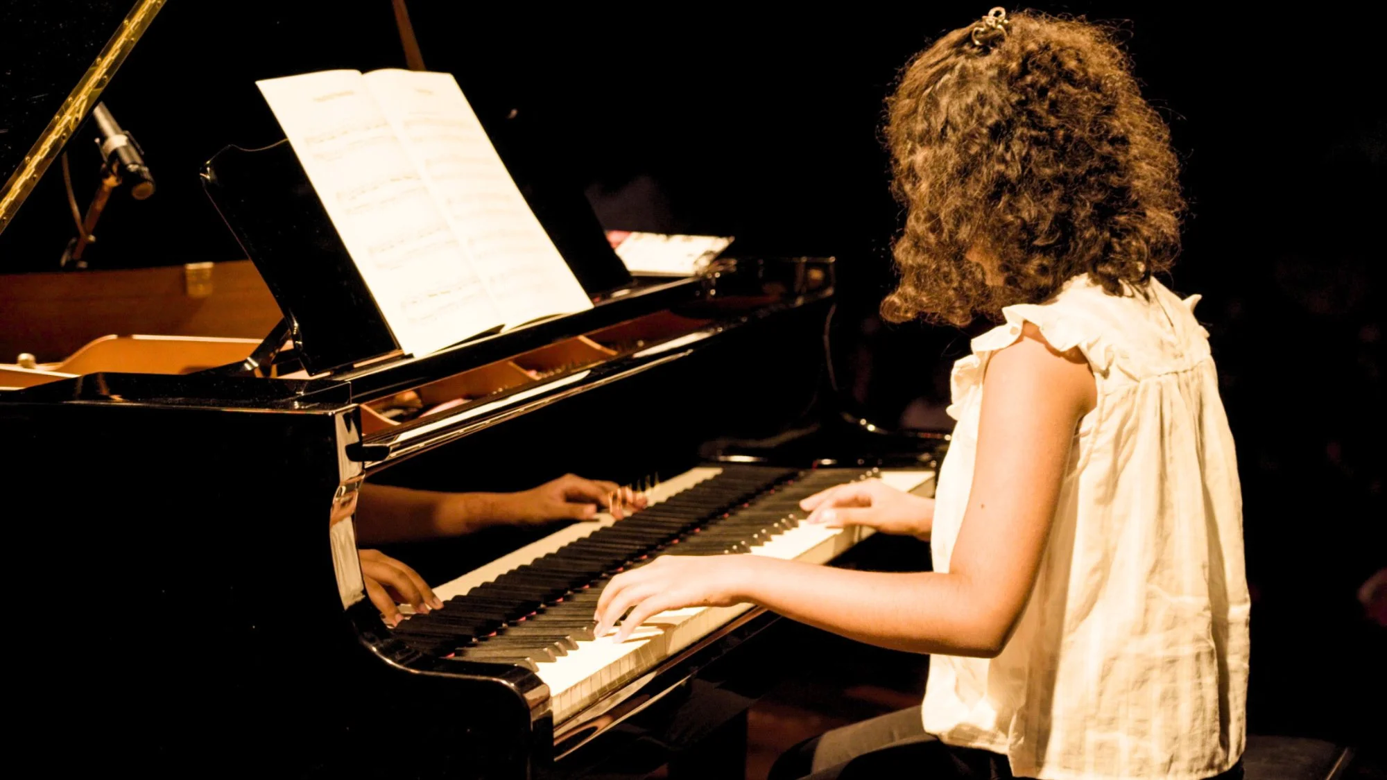 Young girl piano student performing on Kawai grand piano — warm golden stage light